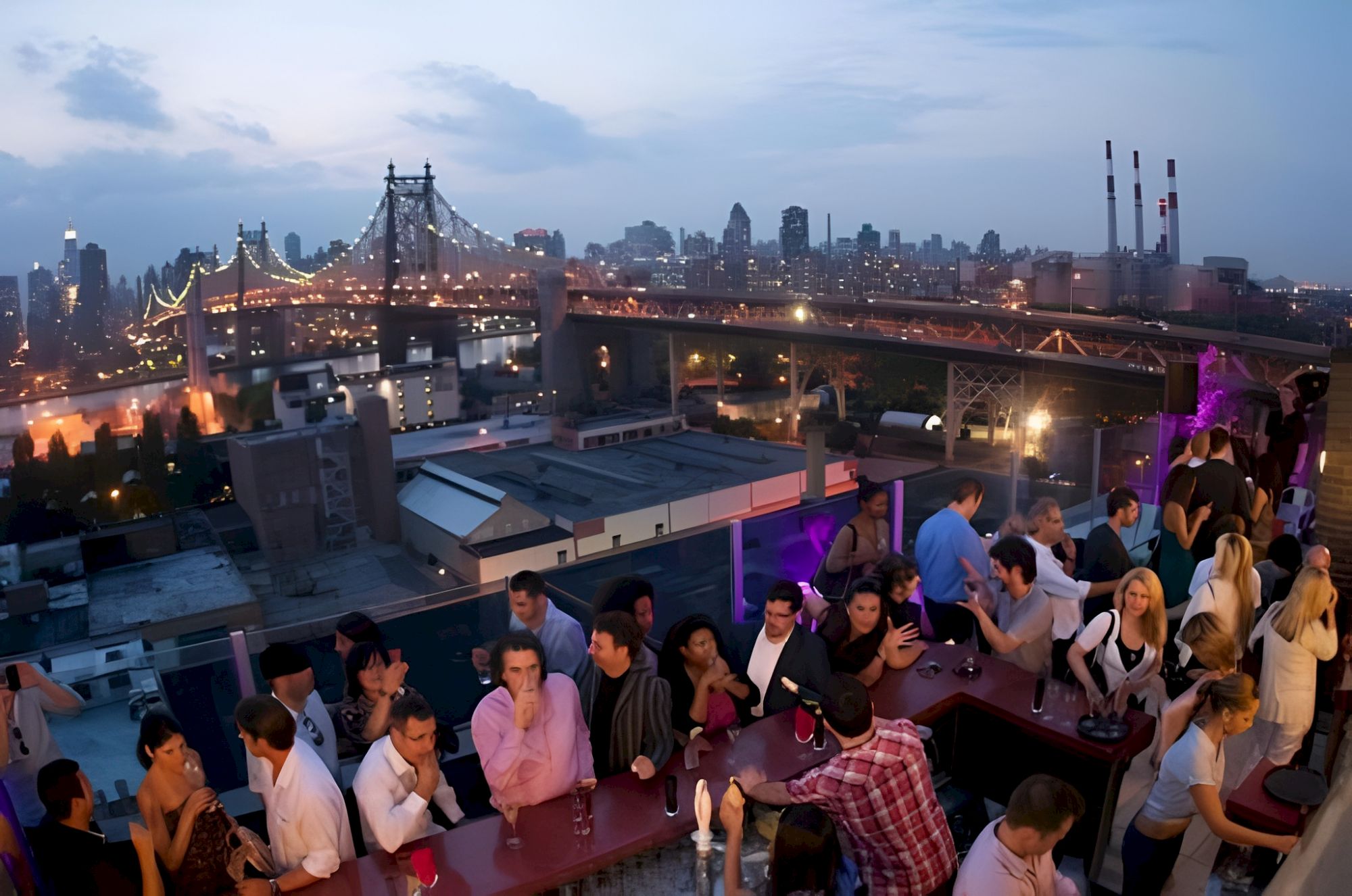 People mingle on a rooftop terrace at dusk overlooking the Brooklyn Bridge and a glowing city skyline.