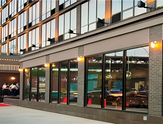Modern building exterior with large glass windows, warm lights, and diners inside, plus a partial storefront sign reading &ldquo;Ho&rdquo;.