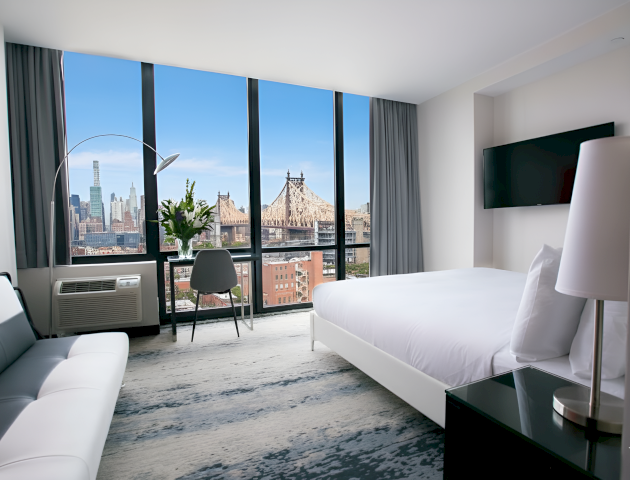 A modern hotel room with a white bed, grey seating, a TV, and large windows showing a city skyline with Brooklyn Bridge.