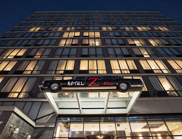 Night view of a tall hotel building as a vintage car marked &ldquo;HOTEL Z NYC&rdquo; sits on a rooftop platform, under stars.
