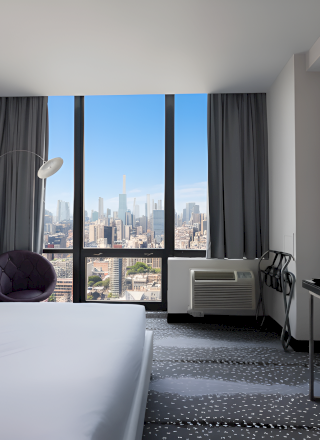 A modern hotel room/office with a bed, desk and chair, a lamp and TV, and a skyline view through large windows.