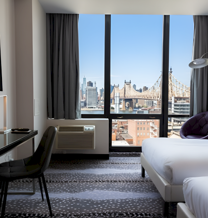 Modern hotel room with a desk and chair, a bed, and floor-to-ceiling windows showing a city skyline and Brooklyn Bridge.