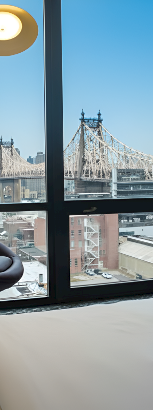 A hotel bedroom with a bed by large windows, looking out at a suspension bridge and city skyline.