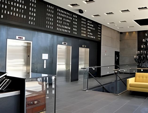 Modern building interior with elevator doors, a dark perforated wall of displays, benches, and polished floors beneath bright ceiling lights.