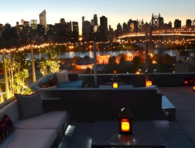 Rooftop lounge at dusk overlooking a glowing NYC skyline and bridge, with city lights reflecting on the river.