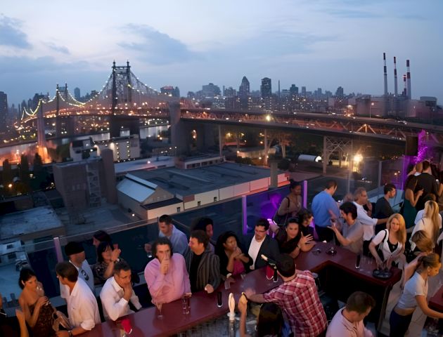 Rooftop party overlooks a lit suspension bridge and a sprawling city skyline at dusk.