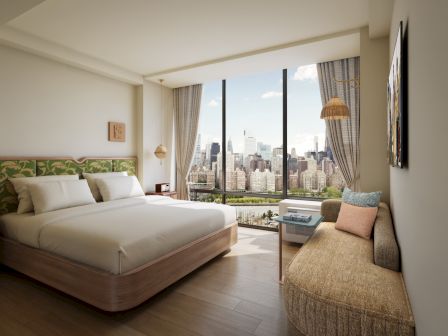 A modern hotel bedroom with a large bed, beige curtains, and a skyline city view through floor-to-ceiling windows.
