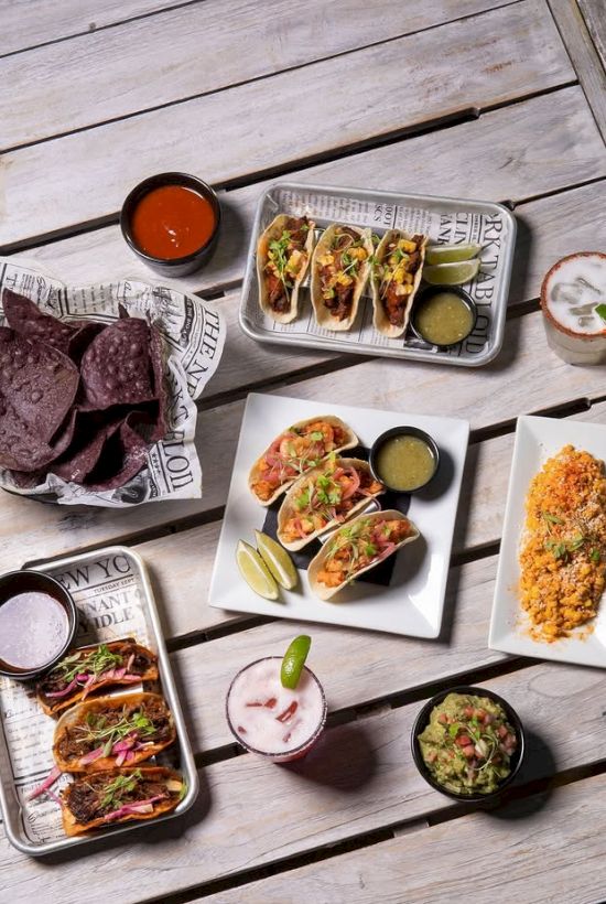 A spread of tacos and small plates with sauces, lime and guacamole on a wooden table.
