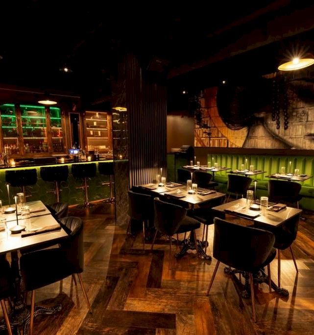 A dimly lit bar/restaurant interior with neatly set tables, dark chairs, neon-green lighting, and rugged industrial decor.