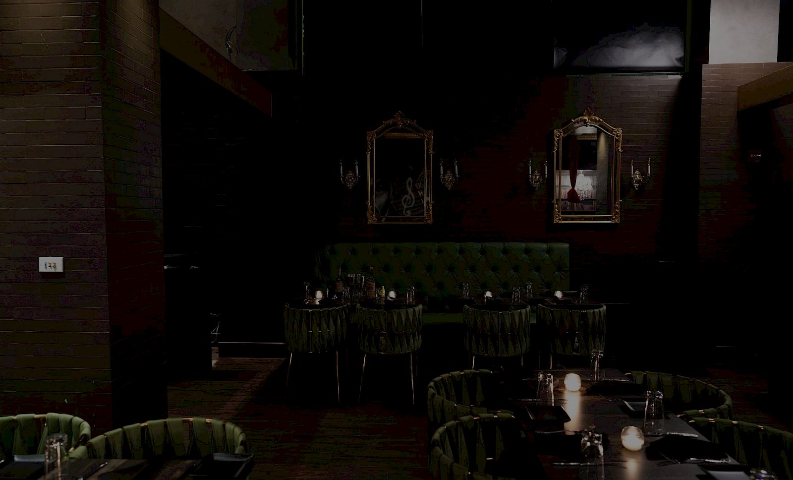 Dimly lit dining room with green upholstered chairs, candles on tables, and framed mirrors on dark walls. Quiet, intimate restaurant setting.